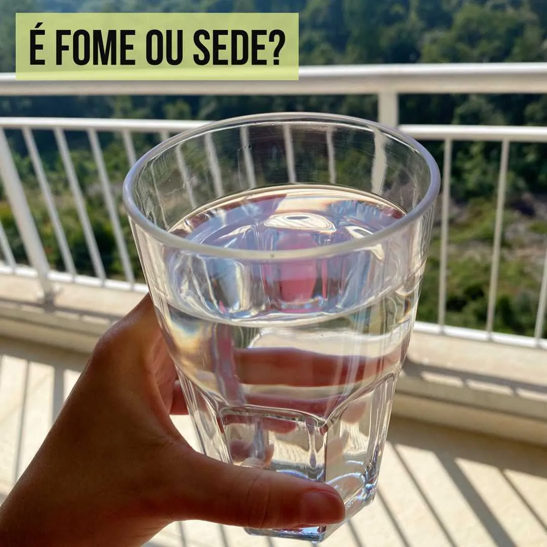 Mão segurando copo de vidro com água numa varanda, lembrando que muita fome é, na verdade, sede