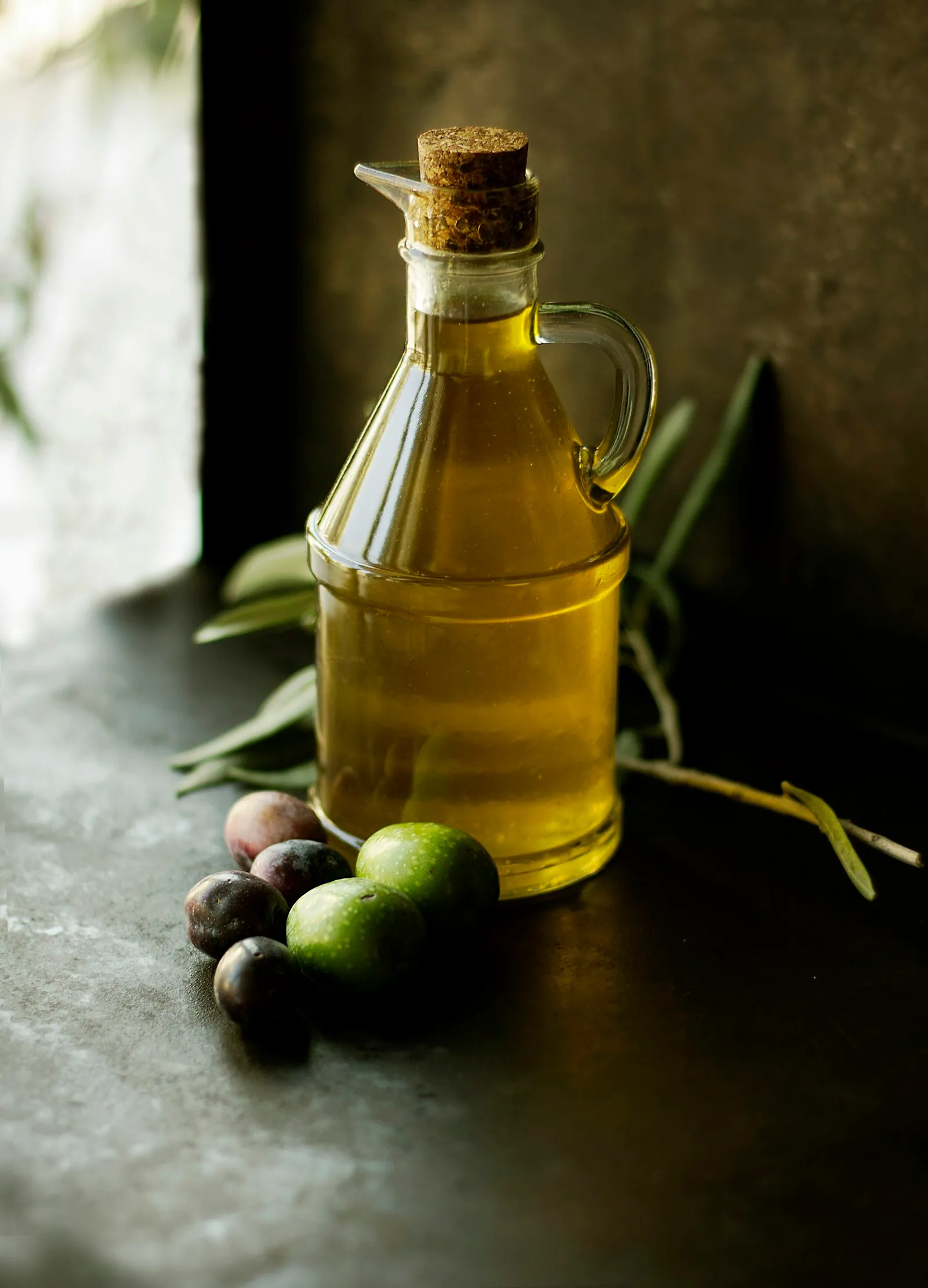 Galheteiro de vidro com azeite de oliva extra virgem e azeitonas verdes e roxas ao lado, sobre bancada escura