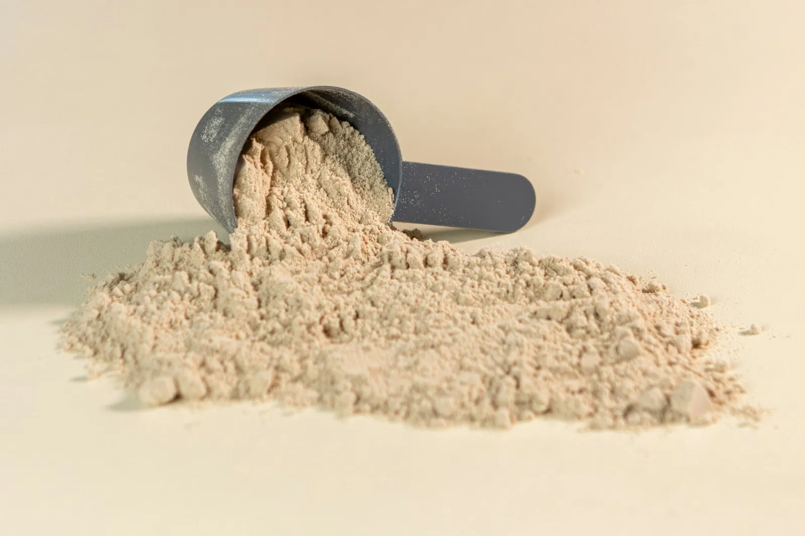 Dosador cinza derramando whey protein em pó bege sobre superfície clara